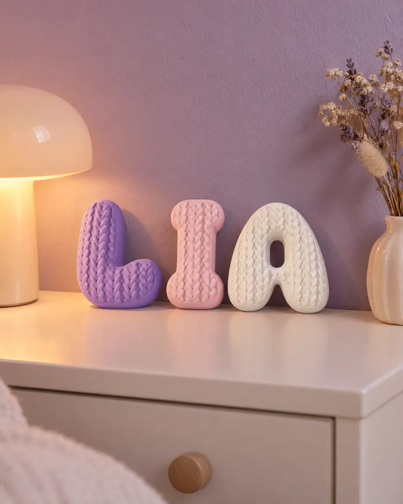 Letras bolha 3D com textura de tricô formando o nome Lia num criado-mudo, ao lado de um abajur cogumelo